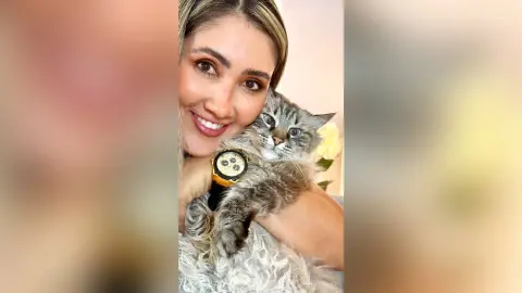 Smiling blonde woman in a white top cuddling a fluffy grey tabby cat in a bright, cozy indoor setting.