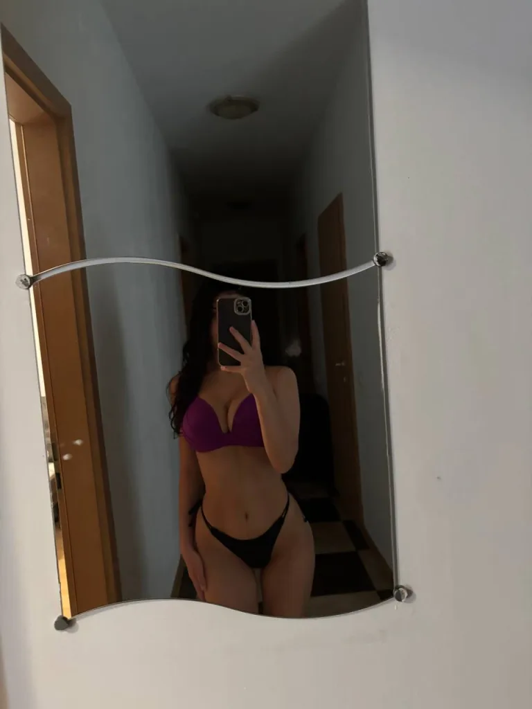 TenderSin poses for a mirror selfie, wearing a white crop top and patterned skirt, showing off her flat stomach and long dark hair.