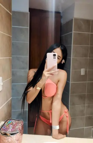 Young woman with long dark hair in a peach bikini taking a mirror selfie in a modern tiled bathroom.