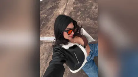 Stylish woman in a black leather jacket and sunglasses posing outdoors on a sunny day.