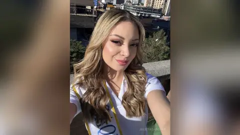 Smiling blonde woman in a white t-shirt posing on a sunny urban balcony with city buildings in the background.