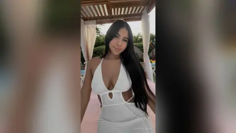 KimberlyIsaza poses in a white cut-out halter dress, showing off her long black hair and cleavage while standing outdoors.