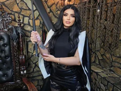 Glamorous woman in a black turtleneck and leather skirt posing with a cane in a moody, stone-walled room.