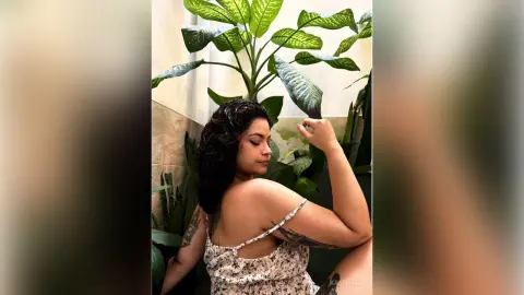 Smiling woman in a floral print sundress posing near lush green tropical plants in a bright indoor setting.