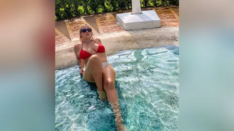 Blonde woman in a red bikini relaxing in a clear turquoise swimming pool on a sunny summer day.