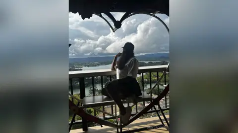 Young woman in a white shirt and black shorts sitting on a balcony overlooking a scenic lake and cloudy sky.