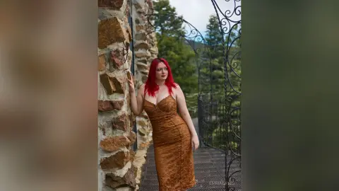 Red-haired woman in a glamorous gold satin dress standing on a stone balcony overlooking a lush forest.