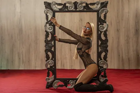 Blonde woman in a black sheer bodysuit posing inside a large ornate vintage frame on a red carpet.