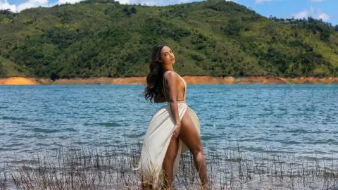 Elegant woman in a white backless maxi dress standing by a serene blue lake with lush green mountains.