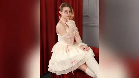 SilvaTailed wears a white ruffled dress with pink bows and sheer gloves, posing in front of red curtains.