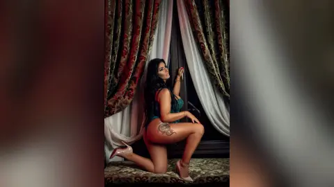 MissyJolie kneels by a window wearing a dark teal lace teddy, showing off her large hip tattoo and long dark hair.