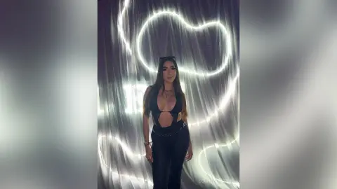 Young woman in a black cutout bodysuit posing against glowing neon heart lights in a moody studio setting.