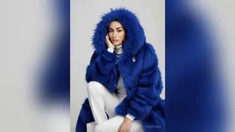 Stylish woman in a cozy blue faux fur hooded coat and white trousers posing against a neutral background.