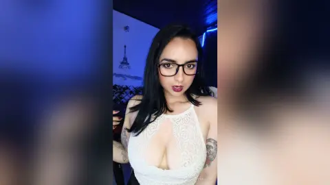Young woman with long dark hair and glasses wearing a white lace top in a moody blue lit room.