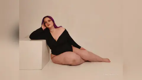 Curvy woman with purple hair in a black v-neck dress sitting on a white pedestal in a minimalist studio.