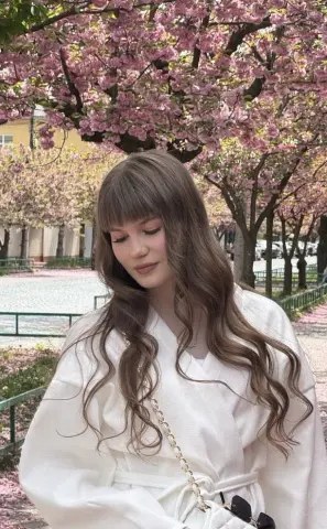 AshleyFierce poses outdoors under pink cherry blossom trees, wearing a white wrap-style robe with her long, wavy brown hair and bangs styled down.