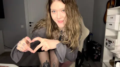 MaryLico smiles while leaning forward, showing her long wavy hair and tattoos on her chest and thighs.