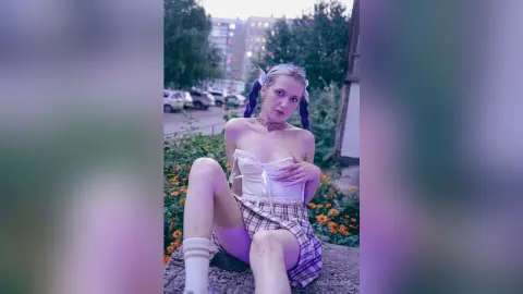 Young woman in a white corset and plaid skirt sitting outdoors in a dreamy, soft-lit urban setting.