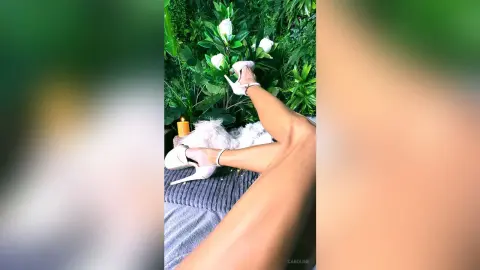 Woman in white high heels relaxing on a cozy grey sofa near lush green plants and a warm candle.