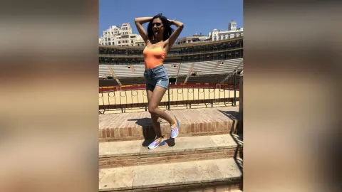 Young woman in orange tank top and denim shorts posing at a sunny outdoor historic amphitheater.