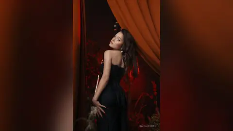 Elegant woman in a black strapless gown posing in a moody, red-lit room with dramatic curtains.