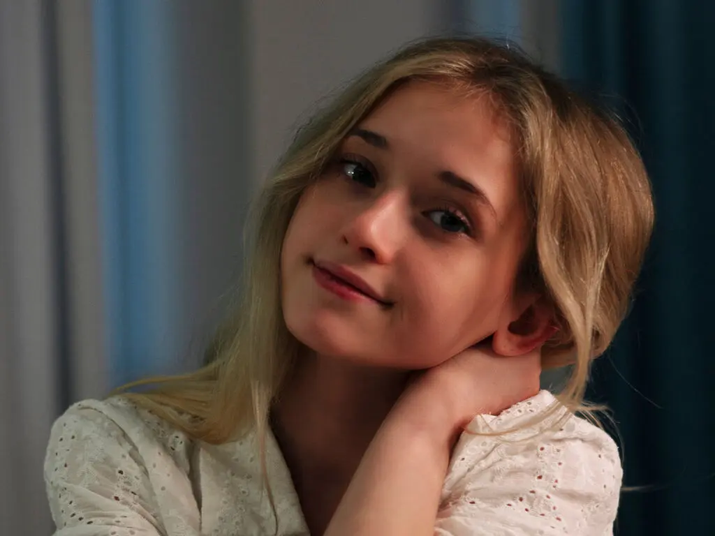 Smiling blonde woman in a white eyelet blouse posing in a soft, moody indoor setting.