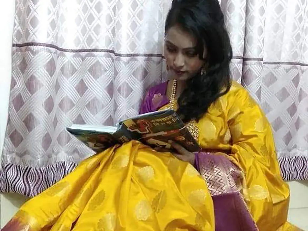 Young woman in a traditional yellow silk saree reading a book in a cozy indoor setting.