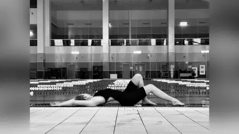 Woman in black bodysuit stretching on a poolside deck in a moody black and white indoor swimming pool setting.