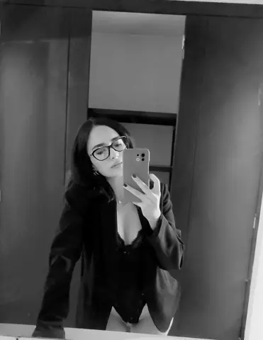 LIAMAXWELL wears black glasses and a dark blazer over a black lace bodysuit while taking a mirror selfie.