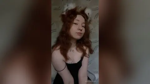 MadgeRemerez poses with red hair and white ears, wearing a black top and a heart necklace against a patterned wall.