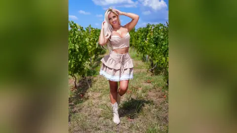 Anita stands in a vineyard wearing a tan lace crop top and matching ruffled skirt with white trim.