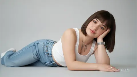 Smiling young woman with short brown hair wearing a white tank top and blue jeans lying on a gray studio floor.