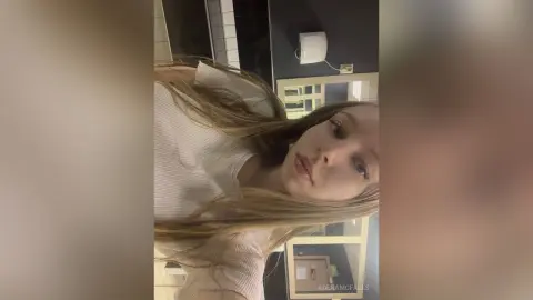 AdenaMcfalls poses close-up with her long blonde hair, wearing a white ribbed tank top or shirt.