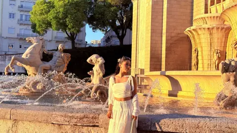 Woman in white summer outfit posing by a classical stone fountain in warm golden hour sunlight.