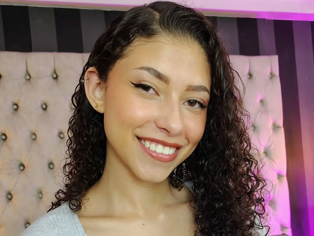 Smiling young woman with curly dark hair and winged eyeliner posing in a cozy bedroom setting.
