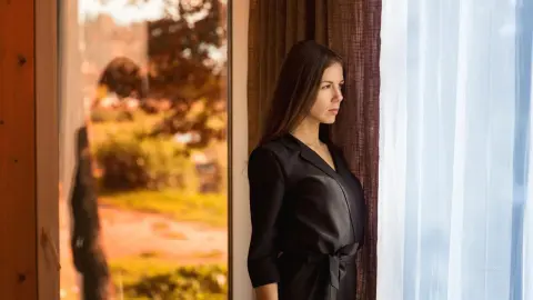 Thoughtful woman in a black satin dress looking out a window in a sunlit room.