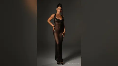 Elegant woman in a sheer black maxi dress posing confidently against a minimalist grey studio background.