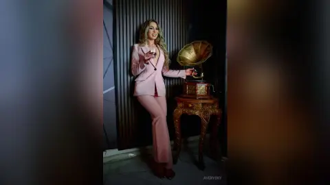 Elegant woman in a pink suit holding a wine glass next to a vintage gold gramophone in a moody interior.