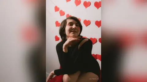 Young woman in a black dress posing happily against a festive red heart garland background.