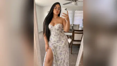 NatalyBloom posing in a mirror wearing a white floral sundress with a high slit, showing off her long dark hair and curves.