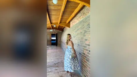 Blonde woman in a blue floral maxi dress standing by a brick wall in a sunlit wooden hallway.