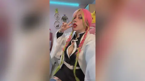 Cosplayer with pink and green hair in a black and white anime outfit posing playfully in a bedroom setting.