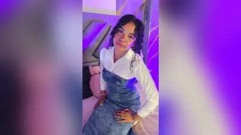 NaomiVelour poses in a white long-sleeve shirt and denim overalls, showing off her curly hair and smile in a purple-lit room.