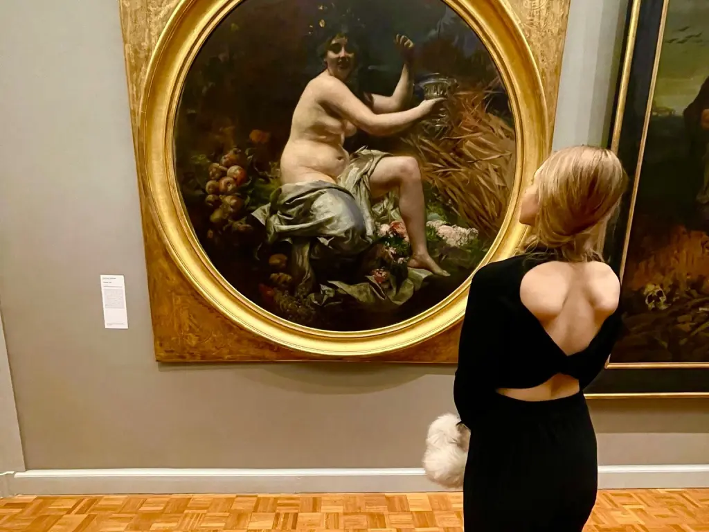 Blonde woman in a black backless dress admiring a classical gold-framed painting in an art museum gallery.