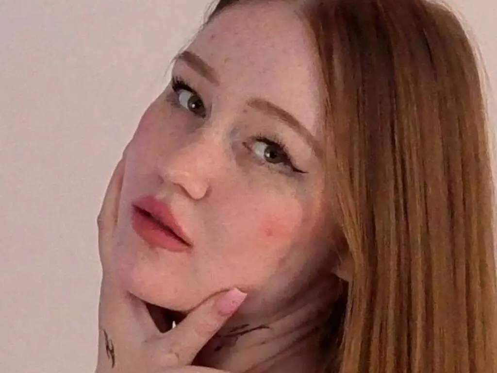AddieRunk poses with her hands on her cheeks, showing her face, freckles, and long reddish-brown hair against a plain background.