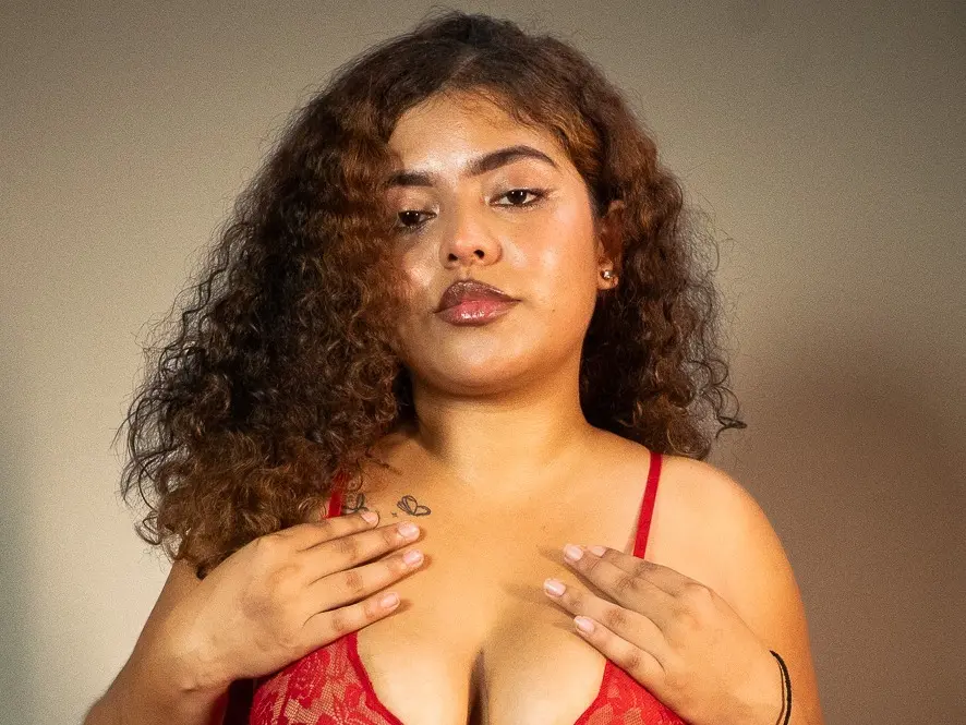 AlanaVelascos poses with her hands on her chest, wearing a red lace bra and showing her cleavage against a plain background.
