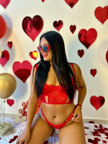 Smiling woman in red lace lingerie posing against a romantic heart-themed backdrop.