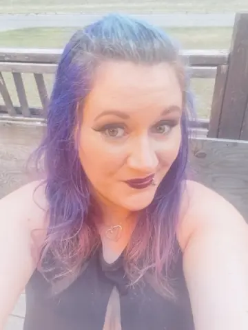 LuxieLuna poses with purple hair and dark lipstick, wearing a black halter-style top while looking directly at the camera.
