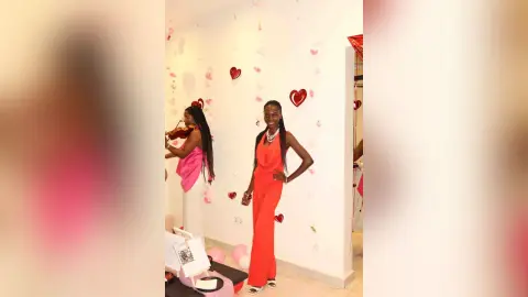 Smiling Black woman in a red jumpsuit posing in a room decorated with red hearts for a romantic celebration.