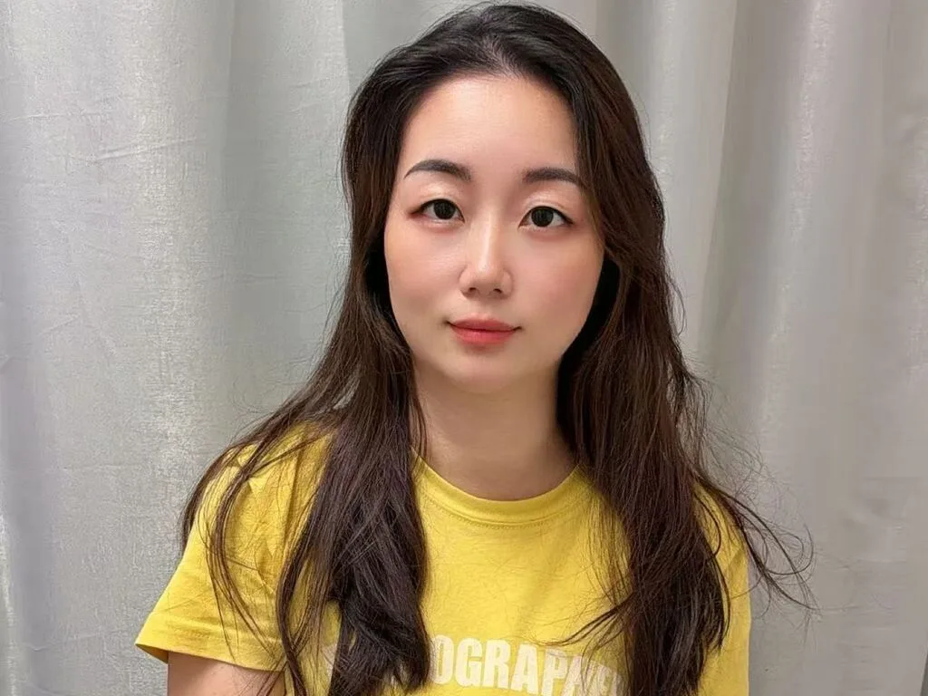 feier looks at the camera wearing a yellow t-shirt with long brown hair against a white curtain background.